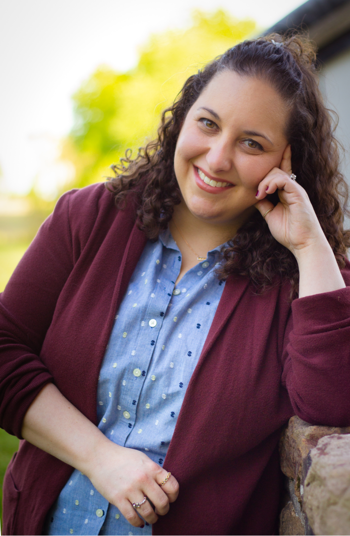 A three-quarter image of Carli J Corson smiling and leaning against a stone fence